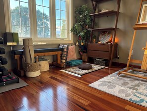 Una habitación con piso de madera, un banco, un estante con adornos y una planta, y un ventanal con vistas a árboles.