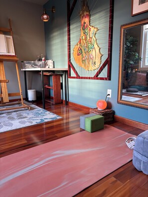 A room with a wooden floor, a yoga mat, a green stool, a painting on the wall, and a ladder.