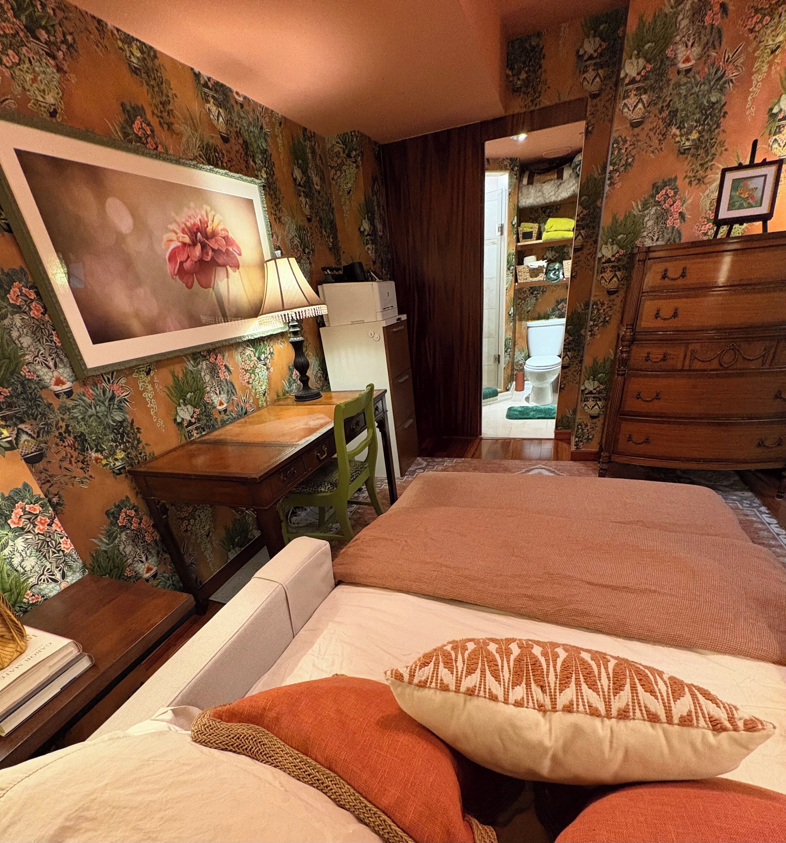 A bedroom with floral wallpaper, a bed with two pillows, a desk with a lamp, and a dresser.