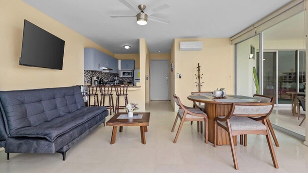 Ein modernes Wohnzimmer mit Sofa, Couchtisch, Esstisch mit Stühlen, einer Küche im Hintergrund und einem Deckenventilator.