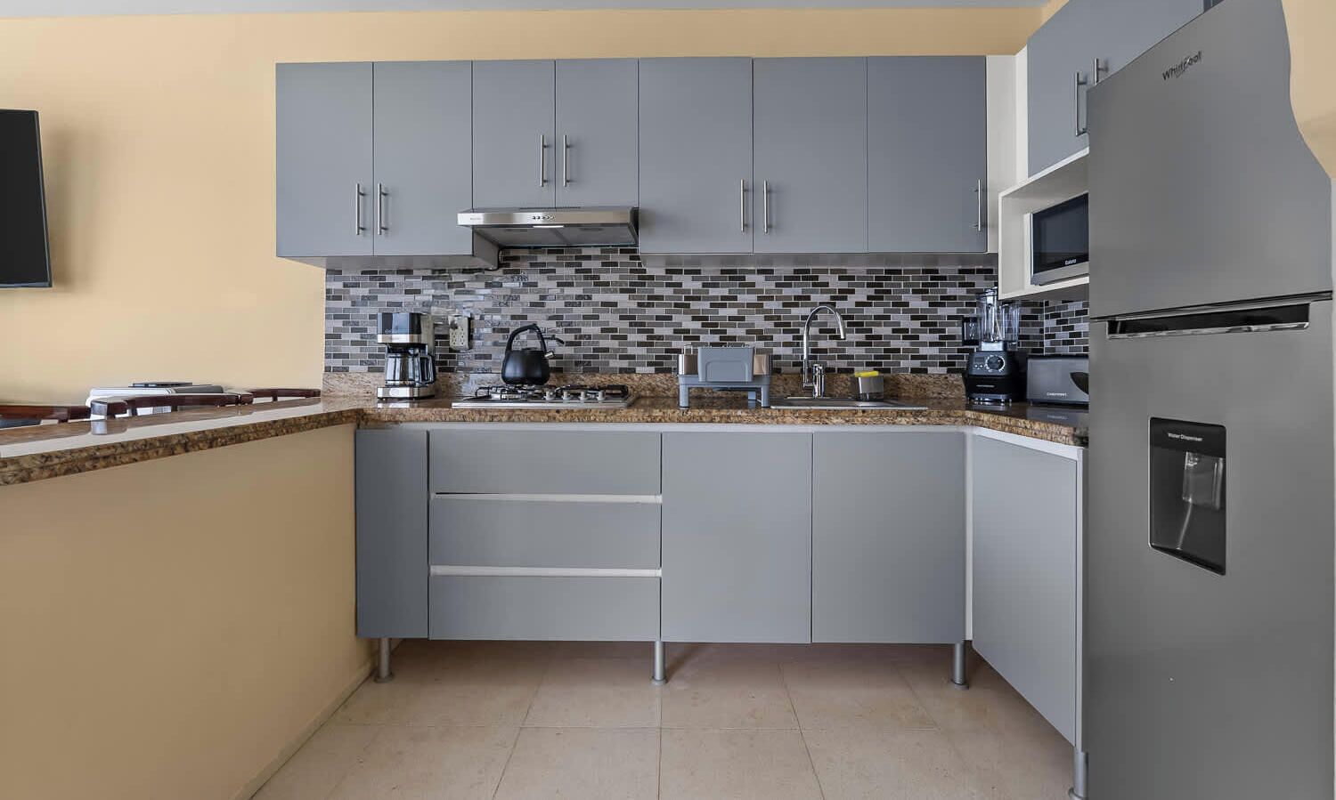 Eine moderne Küche mit grauen Schränken, einer gefliesten Spritzschutzwand und Edelstahlgeräten, darunter ein Kühlschrank und eine Mikrowelle.