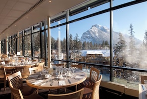 A restaurant with round tables and chairs, large windows offering a view of a snowy mountain. - Banff Centre for Arts and Creativity (Banff)