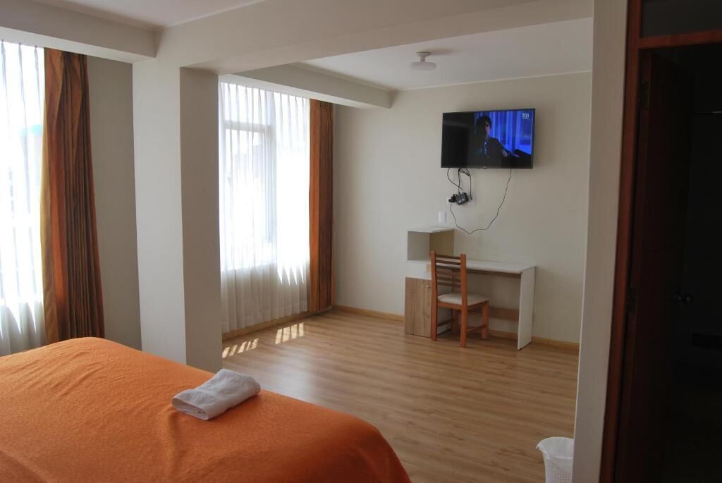 Quarto de hotel com cama, escrivaninha, cadeira, televisão e janela com cortinas.
