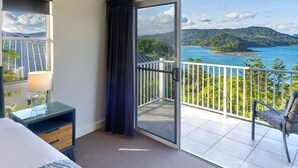 A hotel room with a bed, a nightstand, a lamp, a sliding glass door to a balcony with a view of a beach and mountains. - La Bella Waters 5 and Free buggy - Hamilton Island (Hamilton Island)