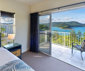 A hotel room with a bed, a nightstand, a lamp, a sliding glass door to a balcony with a view of a beach and mountains. - La Bella Waters 5 and Free buggy - Hamilton Island (Hamilton Island)