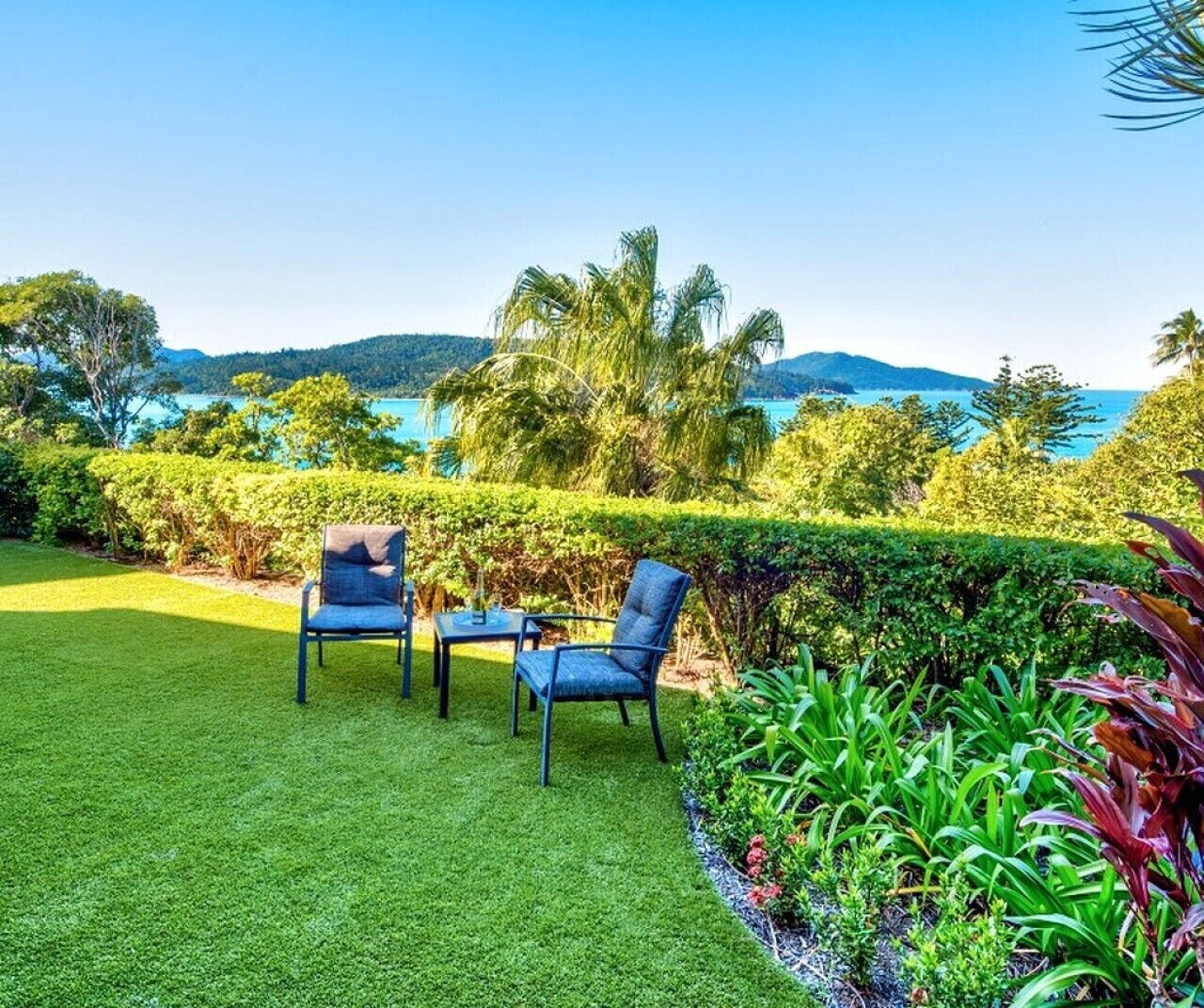 A well-maintained lawn with two chairs and a table, surrounded by lush greenery and a view of the sea.