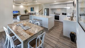 A modern kitchen with a central island, white cabinetry, and a dining area with a rustic wooden table. - La Bella Waters 5 and Free buggy - Hamilton Island (Hamilton Island)