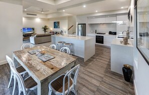A modern kitchen with a central island, white cabinetry, and a dining area with a rustic wooden table. - La Bella Waters 5 and Free buggy - Hamilton Island (Hamilton Island)