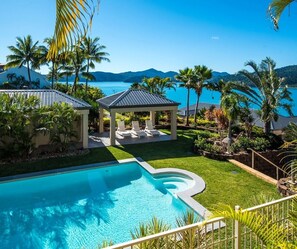 A swimming pool with a curved edge, a covered seating area with lounge chairs, and a view of a body of water and mountains in the background.