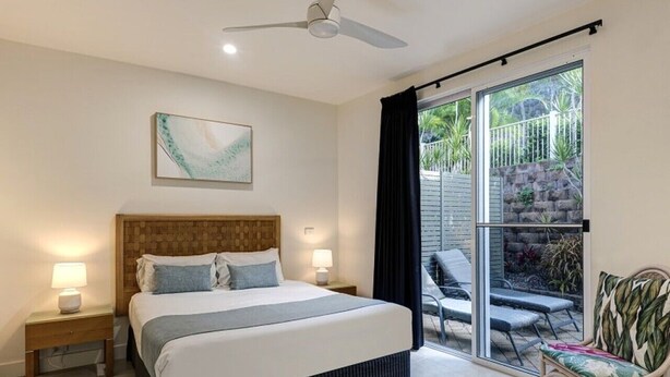 A bedroom with a bed, bedside lamps, a ceiling fan, a large window with curtains, and a view of an outdoor area with chairs and plants. - La Bella Waters 5 and Free buggy - Hamilton Island (Hamilton Island)