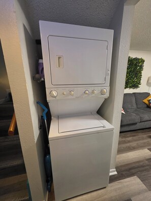 A compact, white laundry room with a built-in washer and dryer, a small shelf with cleaning supplies, and a grey couch in the background. - Waterfront Canal Townhouse with Private Dock on the Island, Corpus Christi (Corpus Christi)