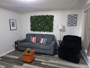 Ein Wohnzimmer mit grauem Sofa, einem schwarzen Sessel, einem kleinen orangefarbenen Tisch mit einer Pflanze und einer grünen Wanddekoration.