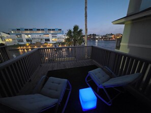 A balcony with two lounge chairs and a blue-lit ottoman, overlooking a waterfront with buildings and a palm tree. - Waterfront Canal Townhouse with Private Dock on the Island, Corpus Christi (Corpus Christi)