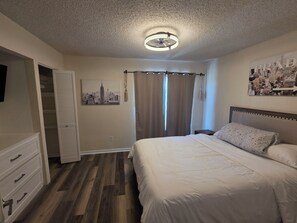 A bedroom with a bed, a nightstand, a dresser, a ceiling fan, a window with curtains, and a cityscape poster on the wall. - Waterfront Canal Townhouse with Private Dock on the Island, Corpus Christi (Corpus Christi)