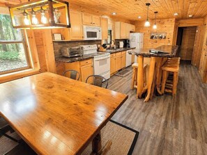 A wooden interior with a central dining table, kitchen area with appliances, and a view through a window. - Log Cabin Sleeps 10 | Pool Pass | Golf + Ski (Bellaire)