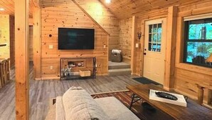 Interior de una cabaña de madera con televisión, sofá, mesa de centro y ventana con vistas.