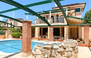 A pool area with a stone waterfall feature, a covered seating area, and a building with a balcony. - Awesome home in Porec (Porec)