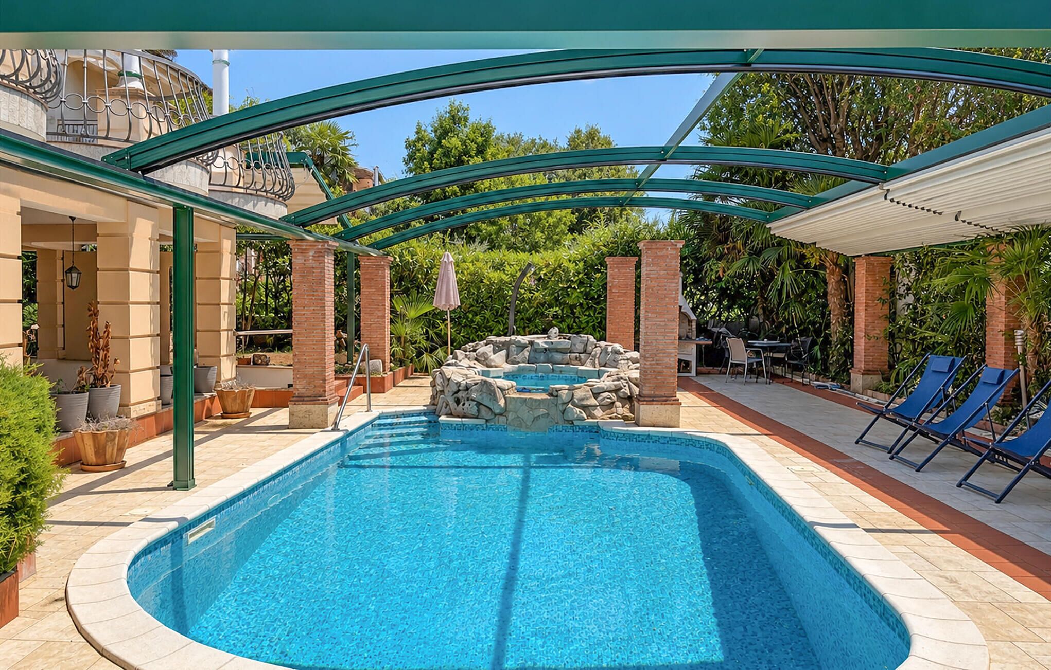 A swimming pool with a stone feature, surrounded by lounge chairs, under a covered patio area.