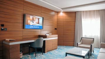 Hotelkamer met een wand van houten panelen, een bureau met stoel, een bank, een salontafel en een televisie waarop een welkomstbericht staat.