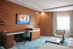 A hotel room with a wooden panel wall, a desk with a chair, a sofa, a coffee table, and a television displaying a welcome message. - Swisse International Rancho Valeria (Warri)