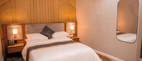 Habitación de hotel con cabecera de madera, un espejo grande, alfombra estampada y mesita de noche con lámpara.