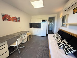 A compact living space with a kitchenette, a desk with a chair, a bed with patterned pillows, and a small rug. - Studio climatisé avec terrasse au calme - La Garde (La Garde)