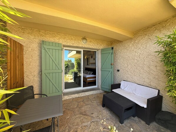 A covered patio area with a table and a sofa, a sliding glass door leading to a bedroom, and a stone wall.
