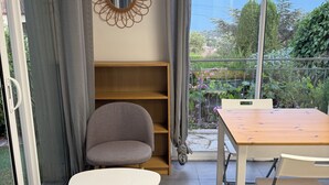 Ein Zimmer mit Esstisch, Stühlen, einem grauen Sessel, einem Holzregal und Blick auf Grün hinter einer Glastür.