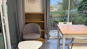 Une pièce comprenant une table à manger, des chaises, un fauteuil gris, une étagère en bois et une vue sur la verdure, visible à travers une porte vitrée.