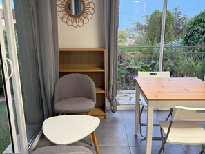 A room with a dining table, chairs, a grey armchair, a wooden shelf, and a view of greenery through a glass door. - Studio cosy dans villa, climatisé, terrasse - La Garde (La Garde)