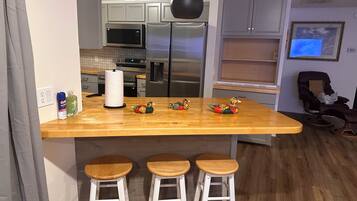 A kitchen with a wooden island, bar stools, a microwave, a refrigerator, and a chair.