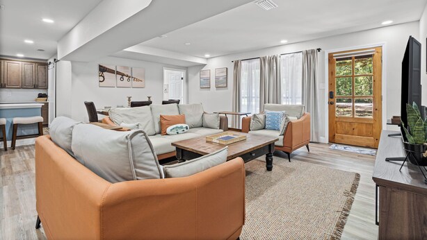 A spacious living area with a sofa, coffee table, and a dining table. There is a kitchen in the background, a large window with curtains, and a wooden door leading outside. - First Cast Foley - Fully Renovated Barn Home (Foley)