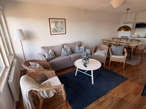Ein Wohnzimmer mit einer Couch, zwei Korbstühlen, einem Couchtisch und einem Essbereich mit Stühlen und Tisch.