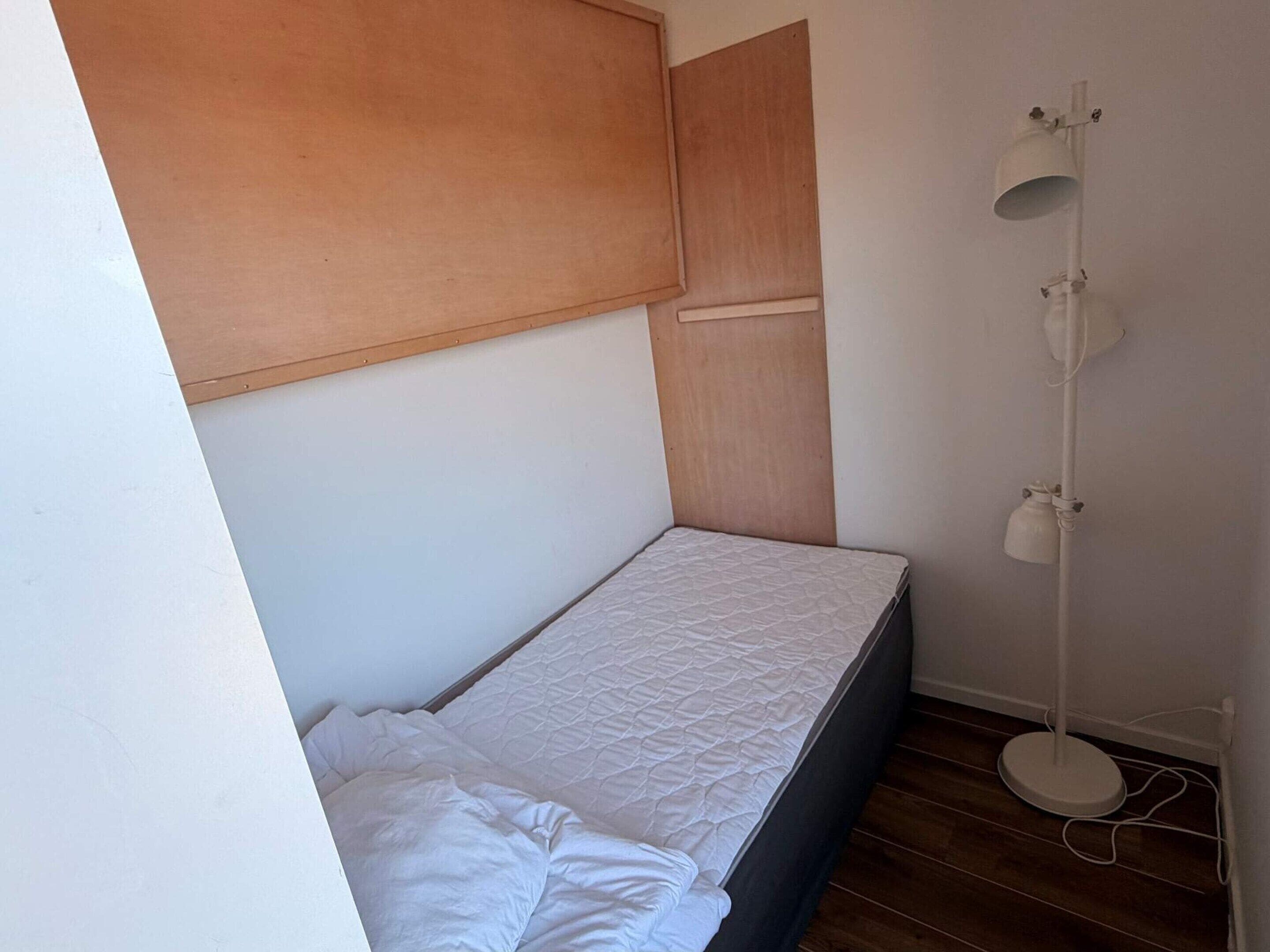 Una habitación pequeña con una cama individual, cabecera de madera, lámpara de noche y suelo de madera.