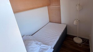Quarto pequeno com uma cama de solteiro, cabeceira de madeira, luminária na mesa de cabeceira e piso de madeira.