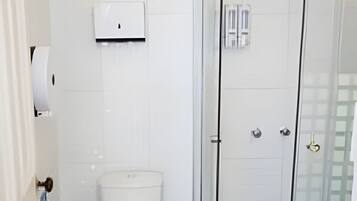 Un baño con inodoro, un cubo de basura, ducha con mampara de vidrio y dispensador de toallas.