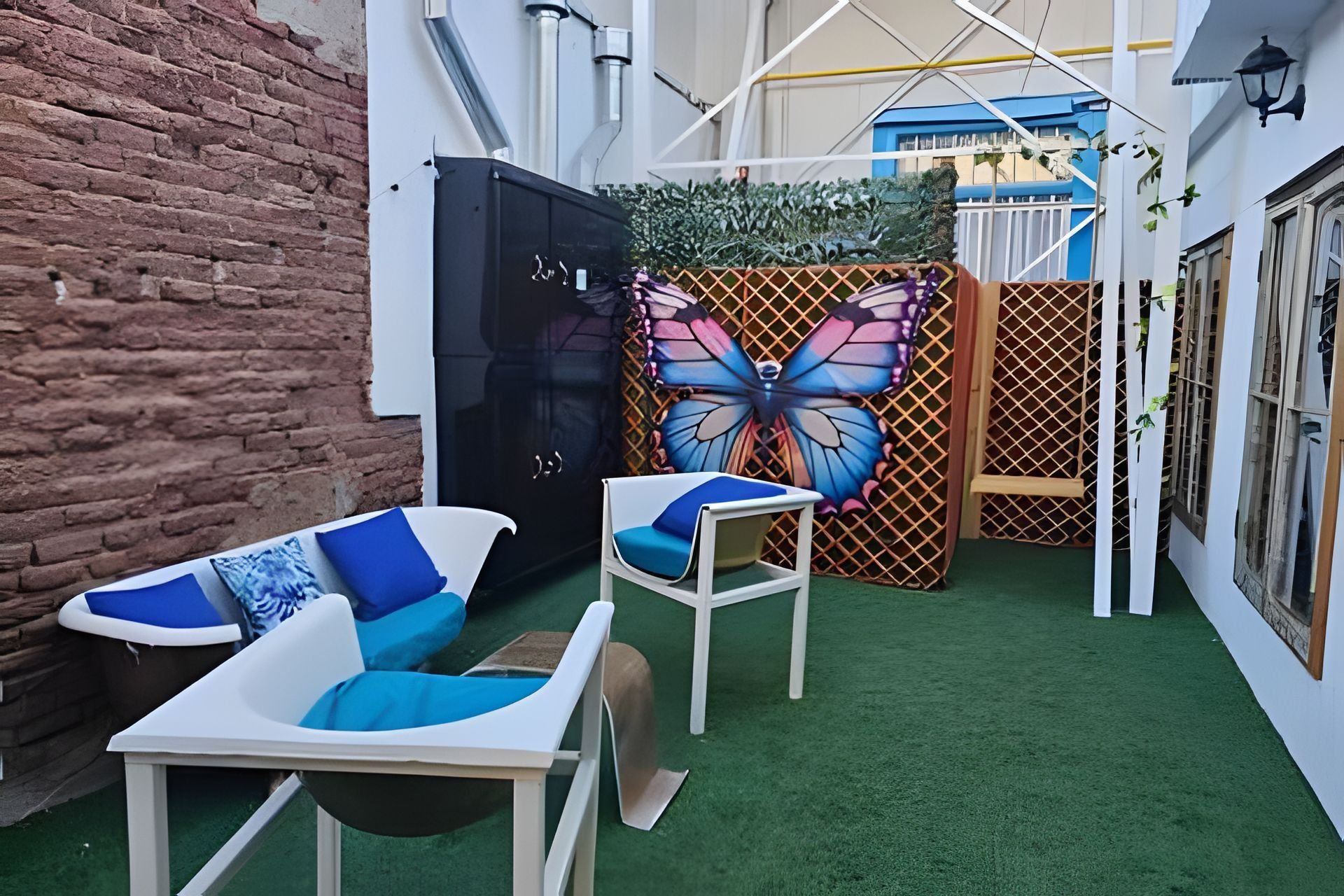 Un patio con pared de ladrillo, un mueble negro, un mural de mariposa y dos sillas blancas con cojines azules.