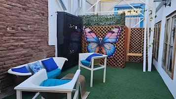 Un patio con pared de ladrillo, un mueble negro, un mural de mariposa y dos sillas blancas con cojines azules.
