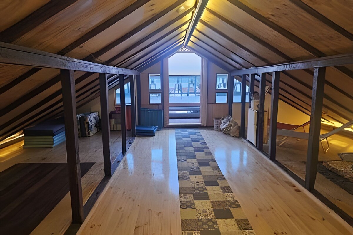 An attic room with a wooden floor, a checkered rug, and a view through a large window.
