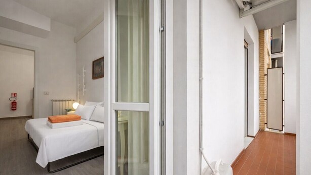 A modern hotel room with a bed, a bedside table, a nightstand, a window with a view, a door, and a hallway leading to a kitchen area. - Cozy Urban Retreat | 2BR | Family Getaway in Rome (Rome)