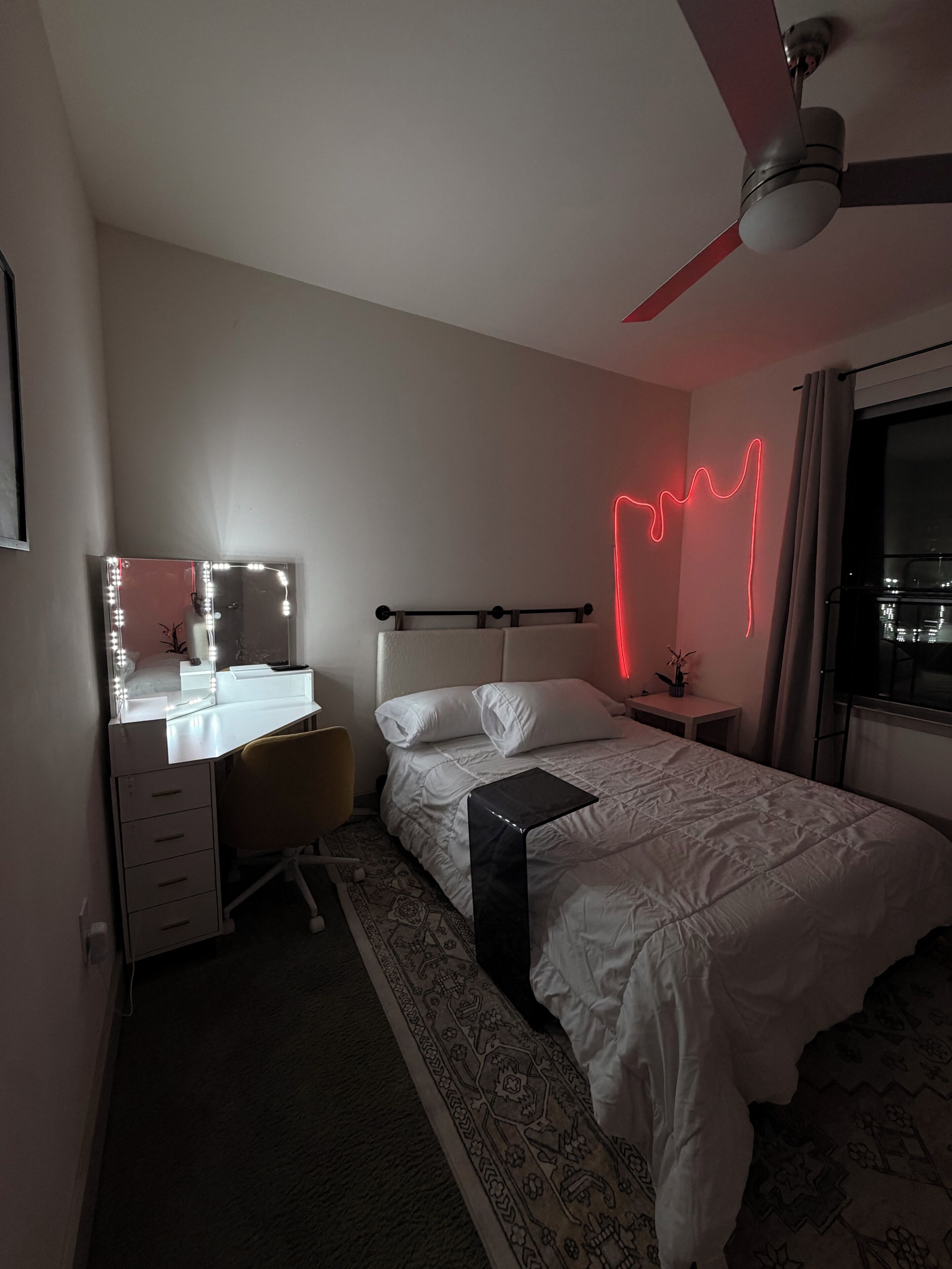 A bedroom with a bed, a desk with a mirror, a ceiling fan, and a nightstand.