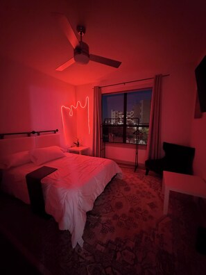 Hotelkamer met een bed, nachtkastje, stoel, raam met uitzicht op gebouwen en een plafondventilator.