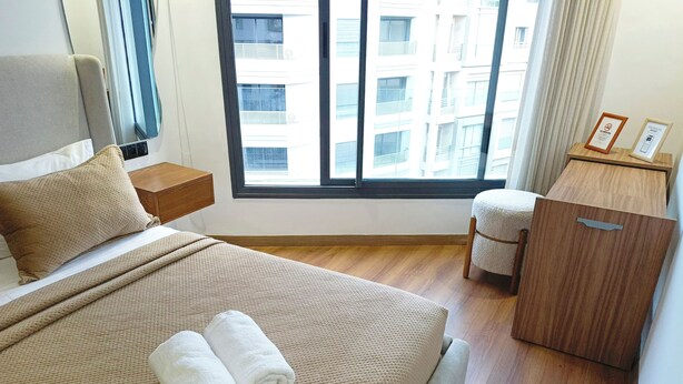 Ein Hotelzimmer mit einem großen Bett, einem Nachttisch aus Holz, einem kleinen Schreibtisch, einem Stuhl und einem Fenster mit Blick auf Gebäude.