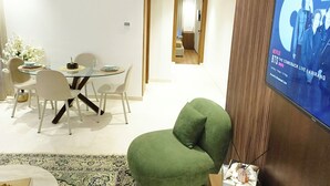 Ein stilvolles Wohnzimmer mit einem Glastisch, weißen Stühlen, einem grünen Sessel und einem Fernseher, der eine Live-Übertragung zeigt.