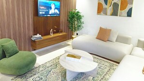 Ein modernes Wohnzimmer mit Flachbildfernseher, einer Couch, einem grünen Sessel, einem Couchtisch und einer Zimmerpflanze.