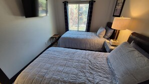 Ein Zimmer mit zwei Betten, einem Nachttisch mit Lampe, einem kleinen Tisch und einem Fenster mit Vorhängen.
