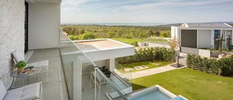 A modern balcony with a glass railing, a pool, and a view of a green landscape.