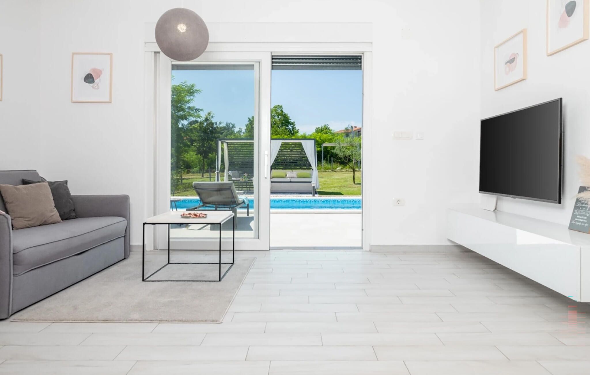A modern living room with a grey sofa, a coffee table, a TV, and a sliding glass door leading to a pool area.