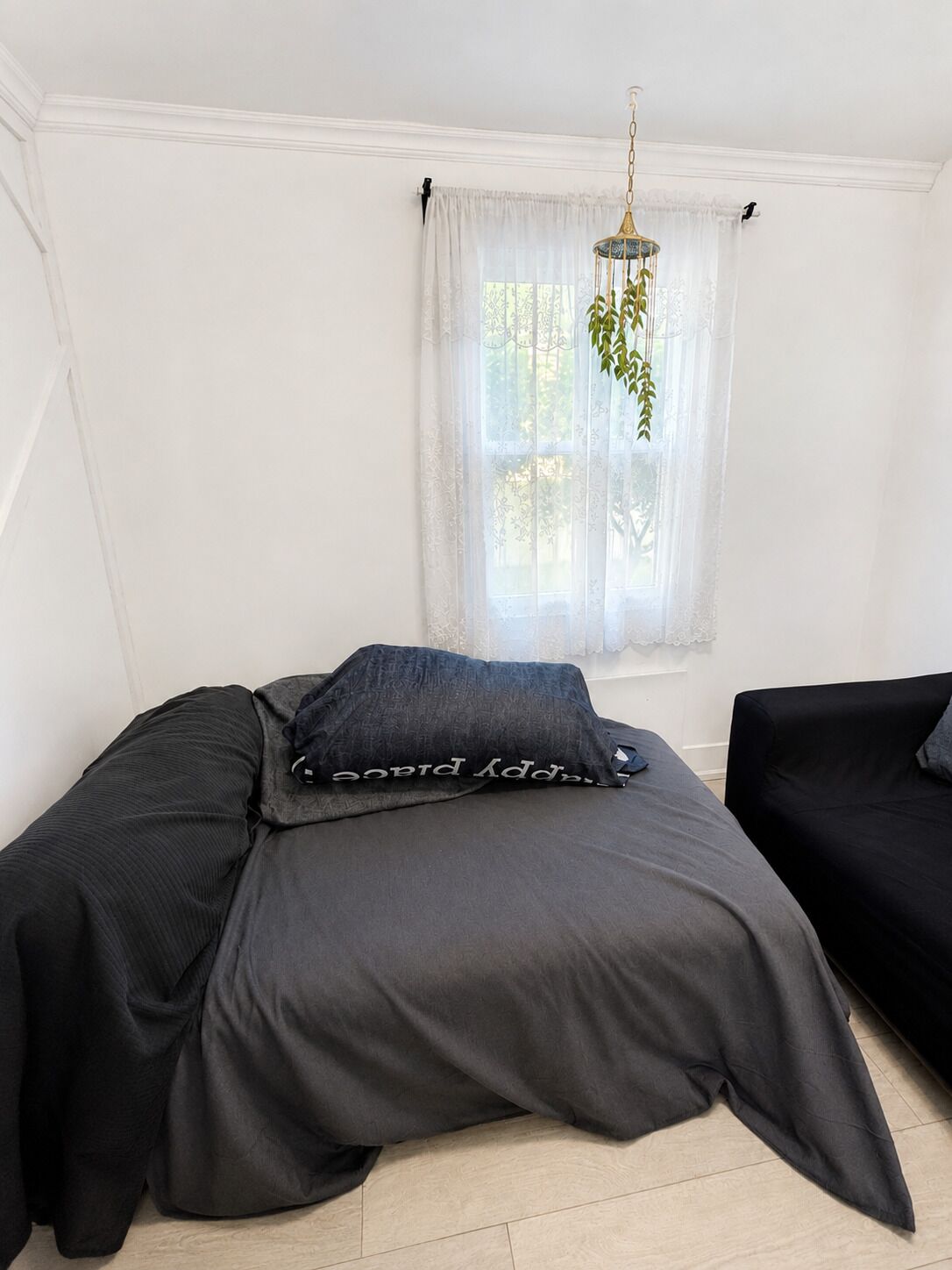 A bedroom with a bed, a hanging plant, and a window with sheer curtains.
