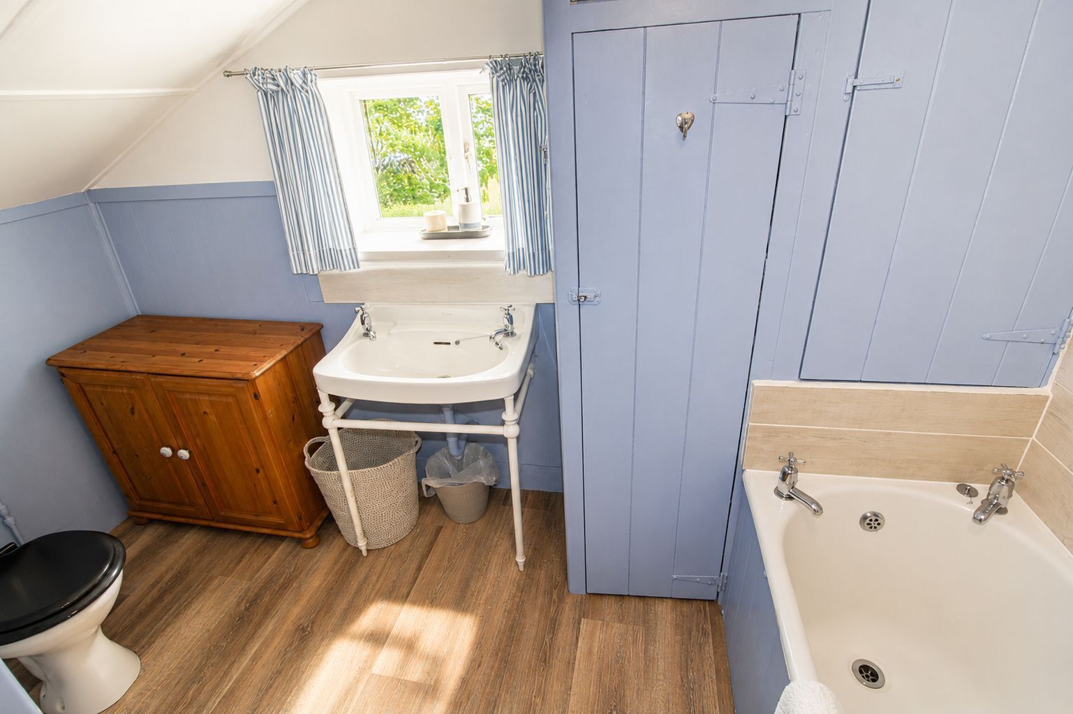 Een kleine badkamer met een houten kastje, een wastafel, een bad en een raam met gordijnen.